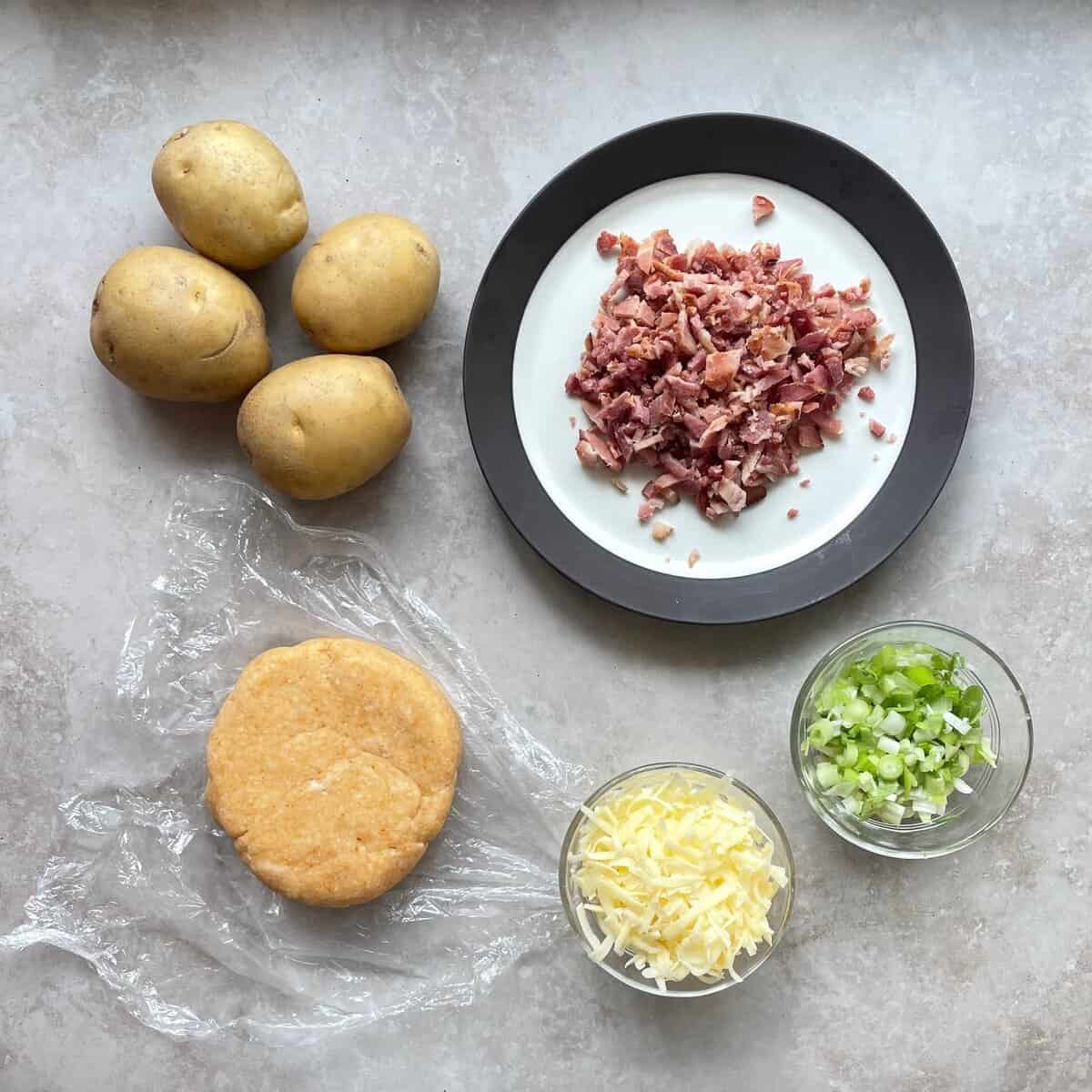 yukon gold potatoes, crisp bacon, green onions, shredded cheddar cheese, and a disc of cheddar cheese pie dough on a countertop.