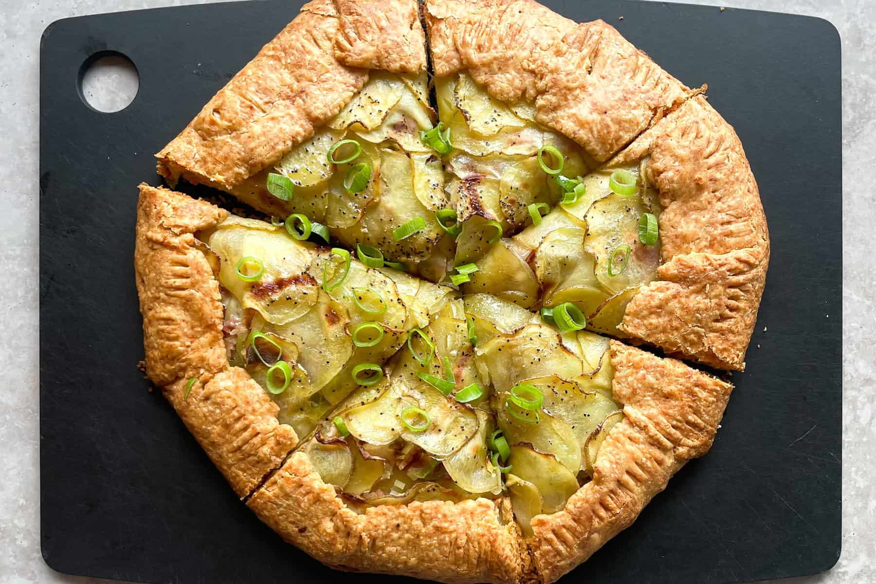 baked potato galette with bacon and green onions cut into six wedges on a cutting board.