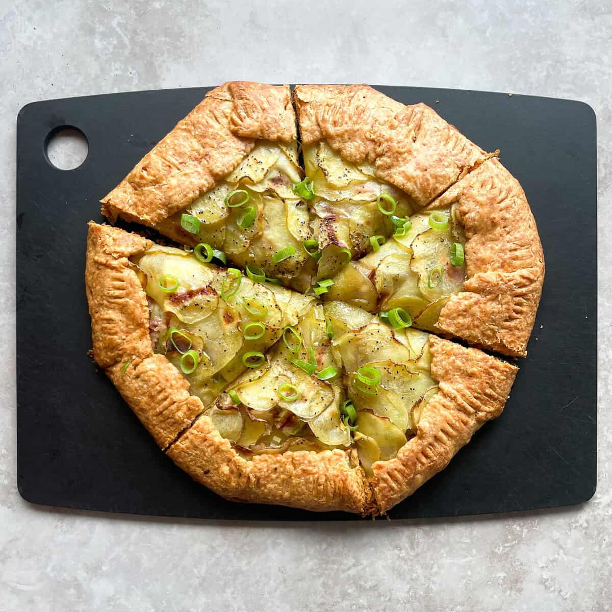 baked potato galette with bacon and green onions cut into six wedges on a cutting board.