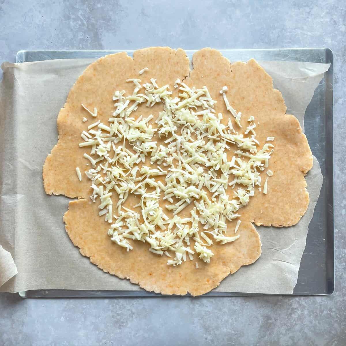 shredded cheddar cheese sprinkled on rolled out cheddar cheese pie dough on a parchment-lined baking sheet.