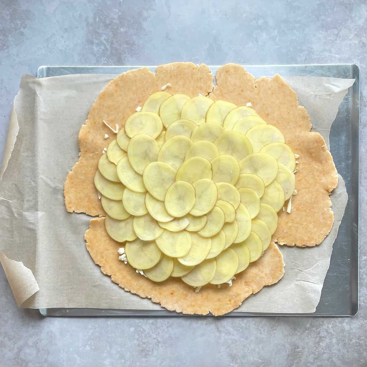 thinly sliced potato layered in overlapping circles on top of the cheddar cheese pie dough on a parchment-lined baking sheet.
