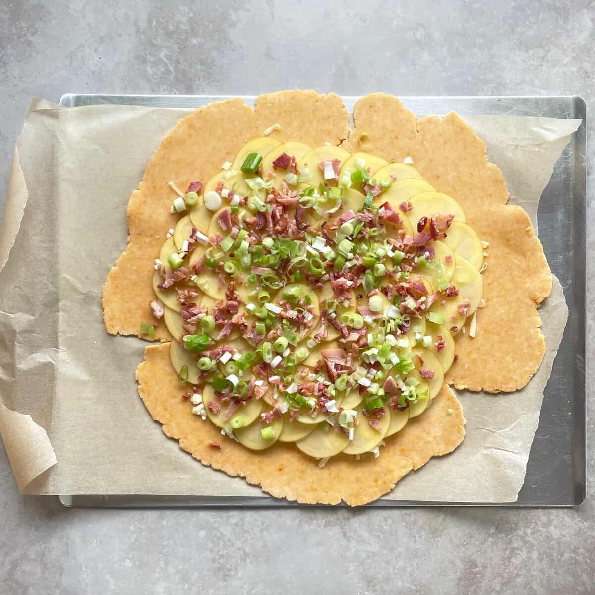 unbaked potato galette with bacon, cheddar cheese, and green onion on top of a layer of potato slices on top of cheese pie dough.
