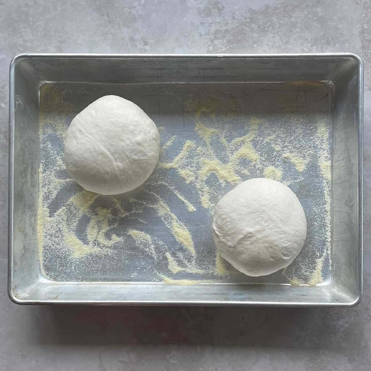 two pizza dough balls in a metal baking pan with semolina flour.