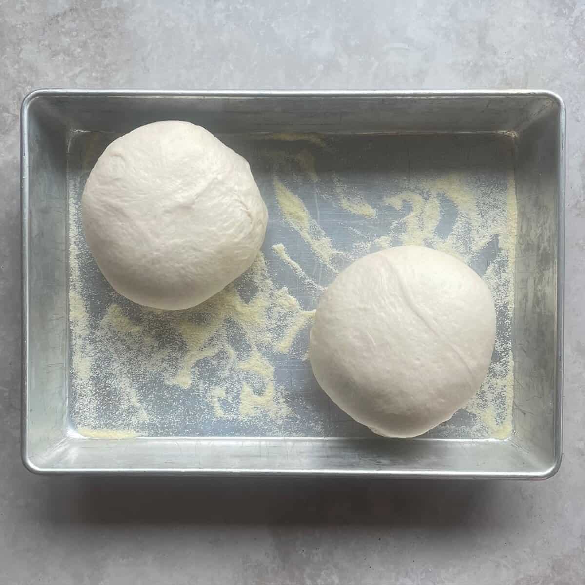 two fully risen pizza dough balls in a metal baking pan with semolina flour.