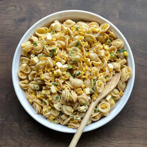 bowl of Mexican street corn pasta salad made with blackened corn kernels, orecchiette, cubes of cheese, and green onion in a creamy dressing with a wooden spoon.