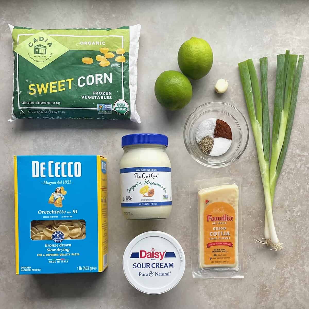 bag of frozen corn kernels, box of orecchiette, limes, mayonnaise, sour cream, cheese, garlic, two green onions, and small bowl of salt, pepper, and other spices.