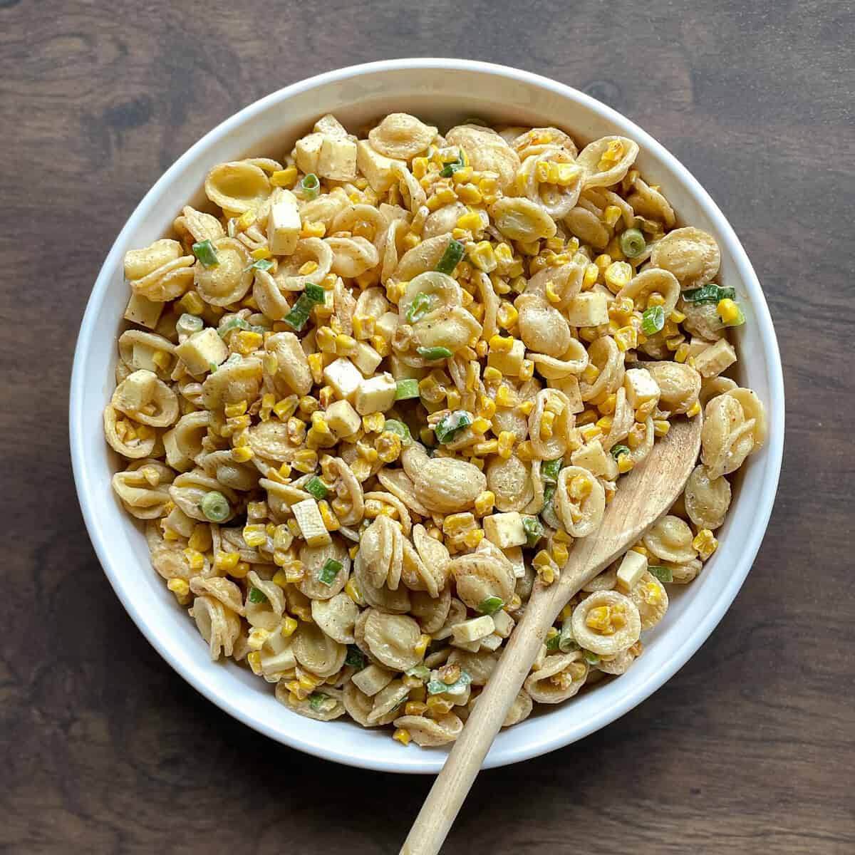 bowl of Mexican street corn pasta salad made with blackened corn kernels, orecchiette, cubes of cheese, and green onion in a creamy dressing with a wooden spoon.