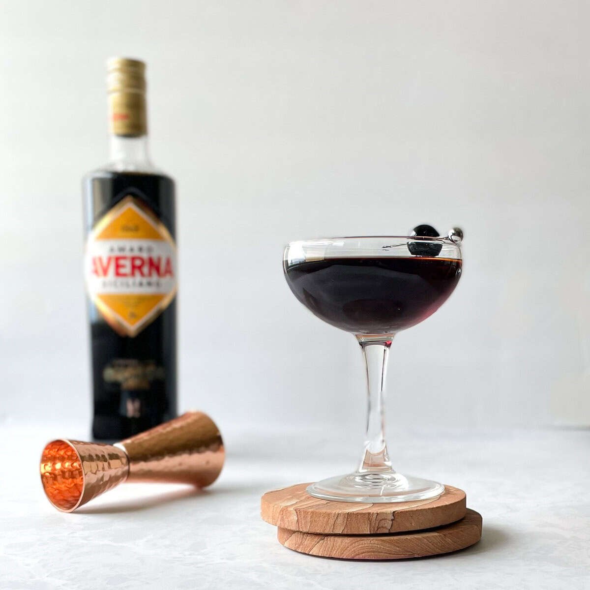 black manhattan and cocktail cherry in coupe next to bottle of averna and copper jigger.