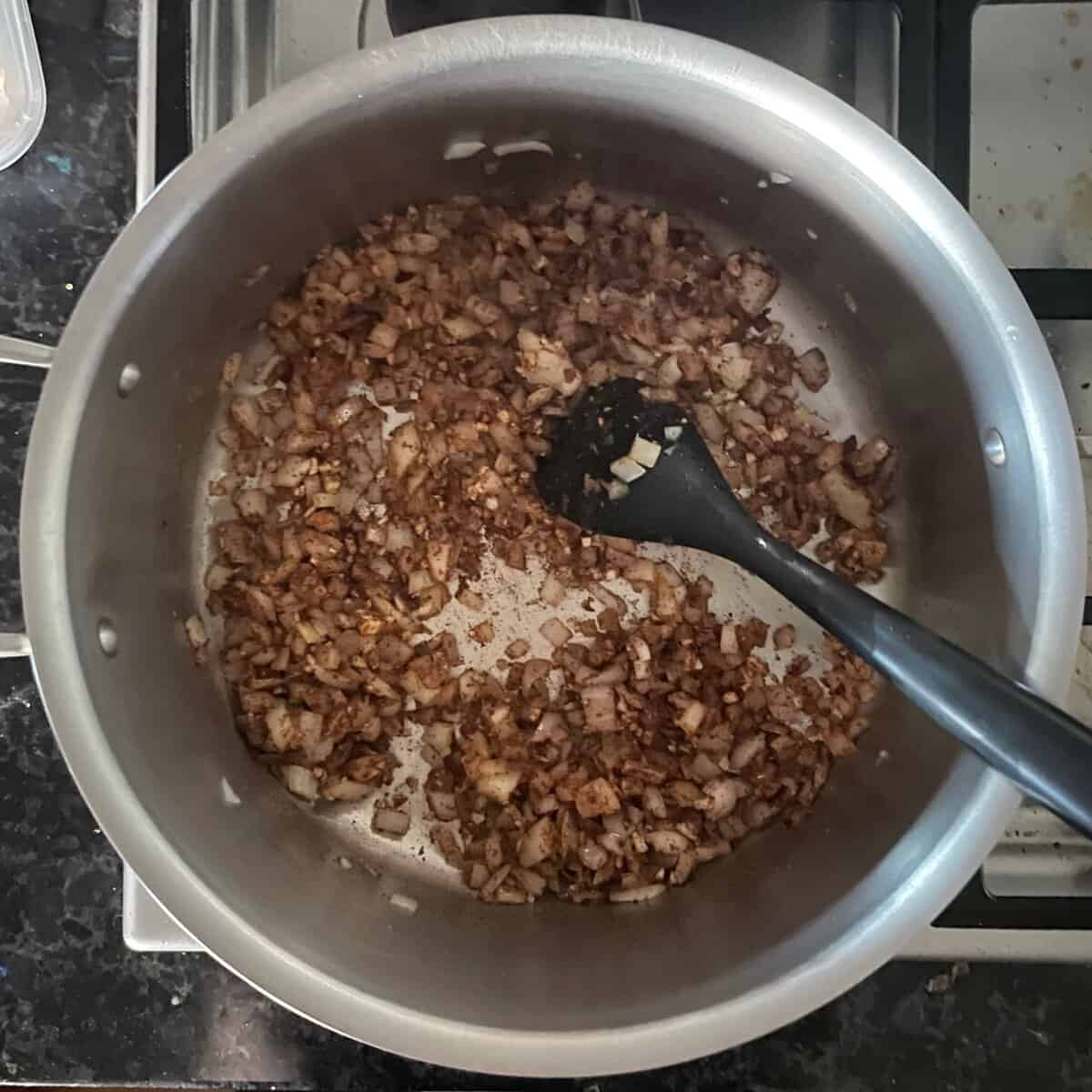 chopped onions, garlic, and toasted spices in a soup pan with black silicone spatula.