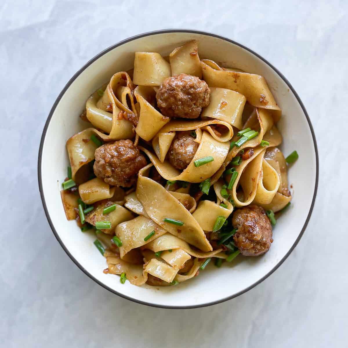pork meatballs in a bowl with wide noodles, green onions, and a brown sauce.