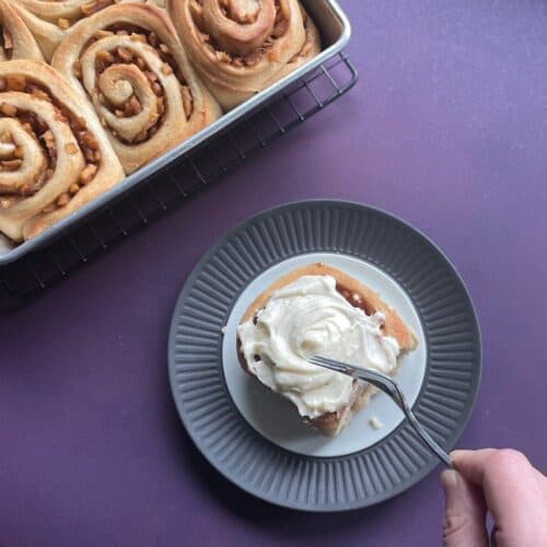 hand holding fork cutting into a frosted apple pie cinnamon roll with pan of apple filled cinnamon rolls in the corner.