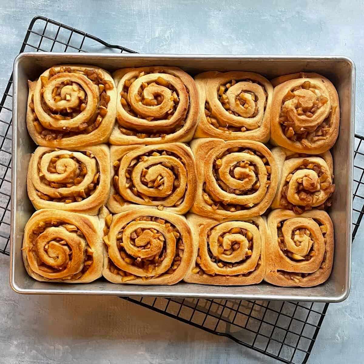 pan of twelve baked apple pie cinnamon rolls on a cooling rack.