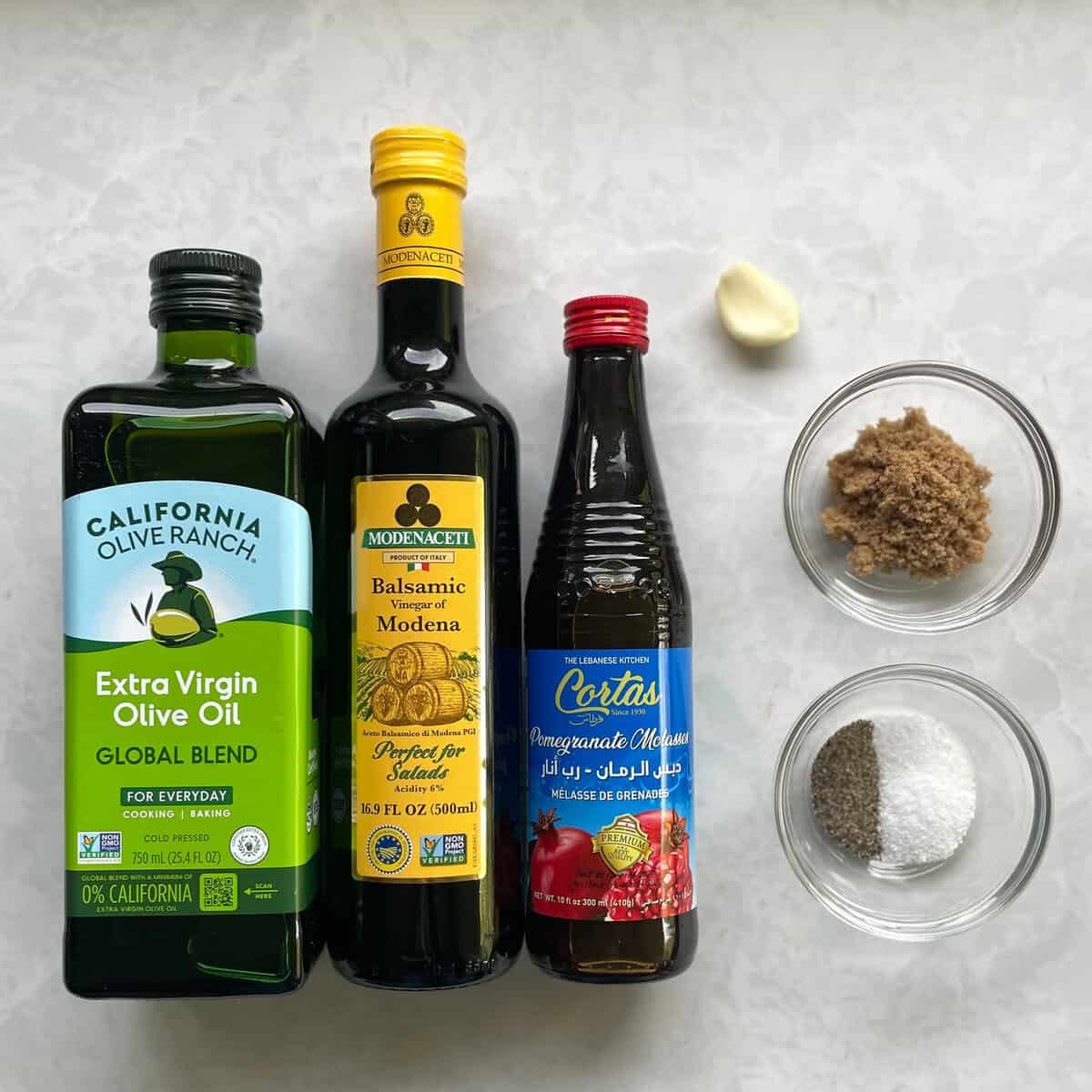 bottles of olive oil, balsamic vinegar, and pomegranate molasses, a garlic clove, brown sugar, and small bowl of salt and pepper.