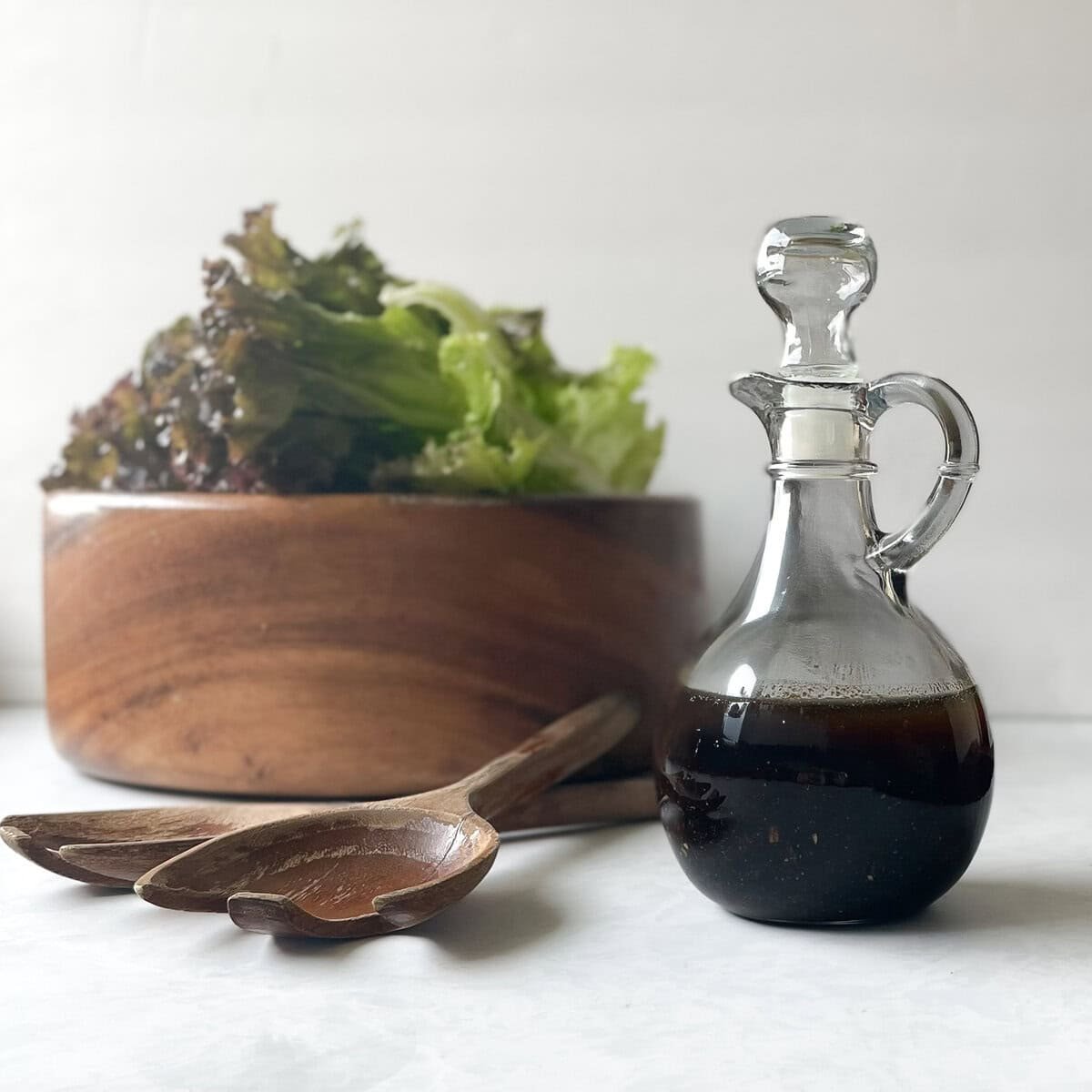 glass jar of salad dressing next to wooden bowl filled with lettuce and wood salad servers.