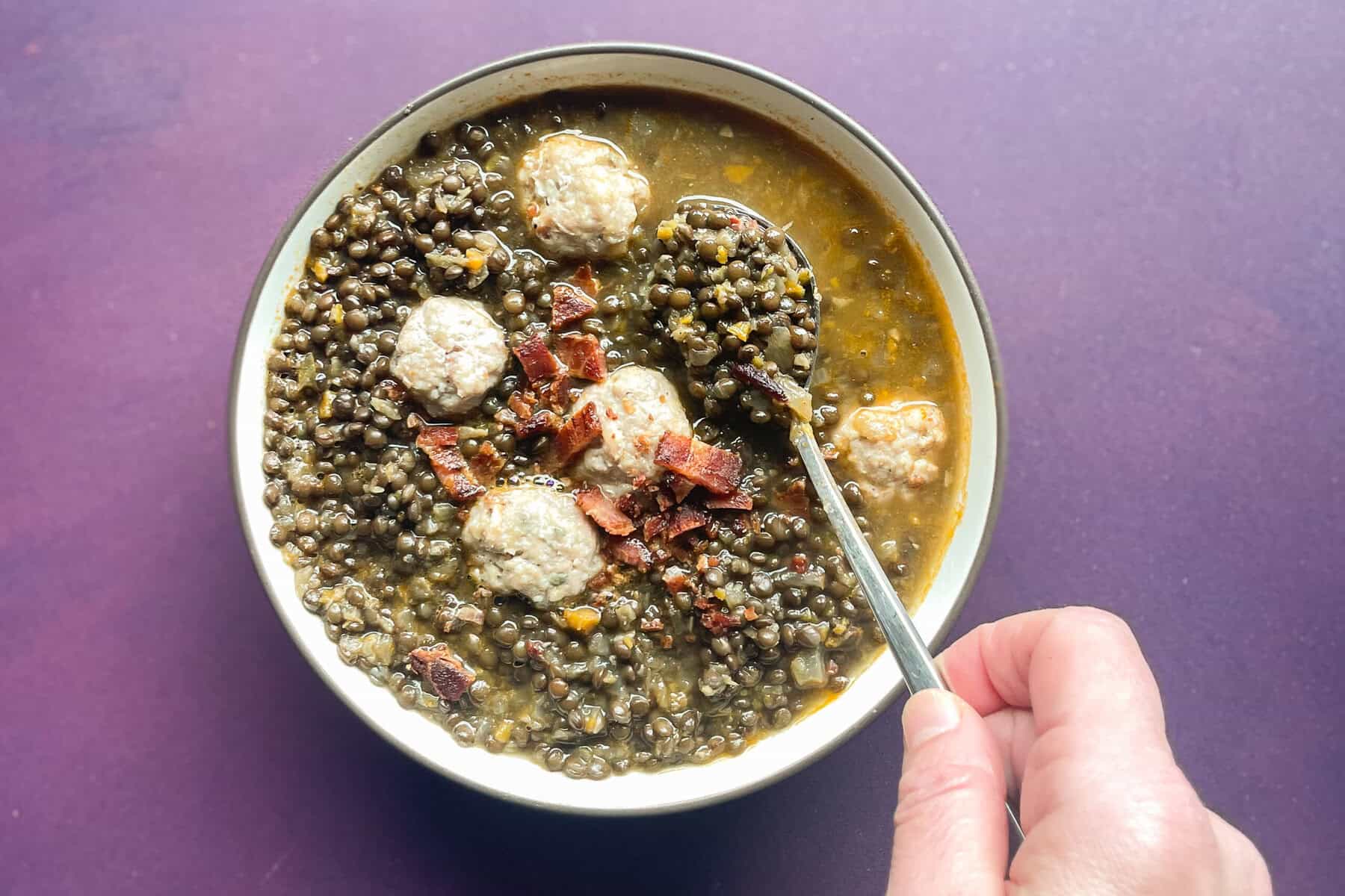 hand holding spoon in bowl of black lentil soup with turkey meatballs and bacon.
