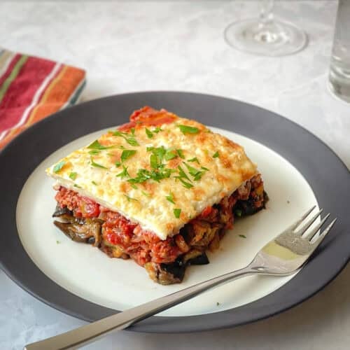 piece of moussaka with layers of eggplant, tomato meat sauce, bechamel, and parsley on a plate with fork.