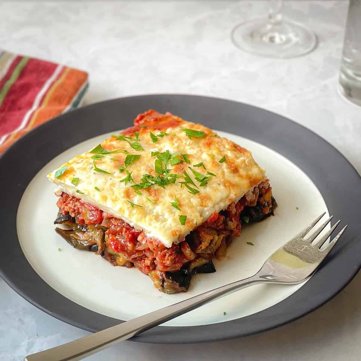 piece of moussaka with layers of eggplant, tomato meat sauce, bechamel, and parsley on a plate with fork.