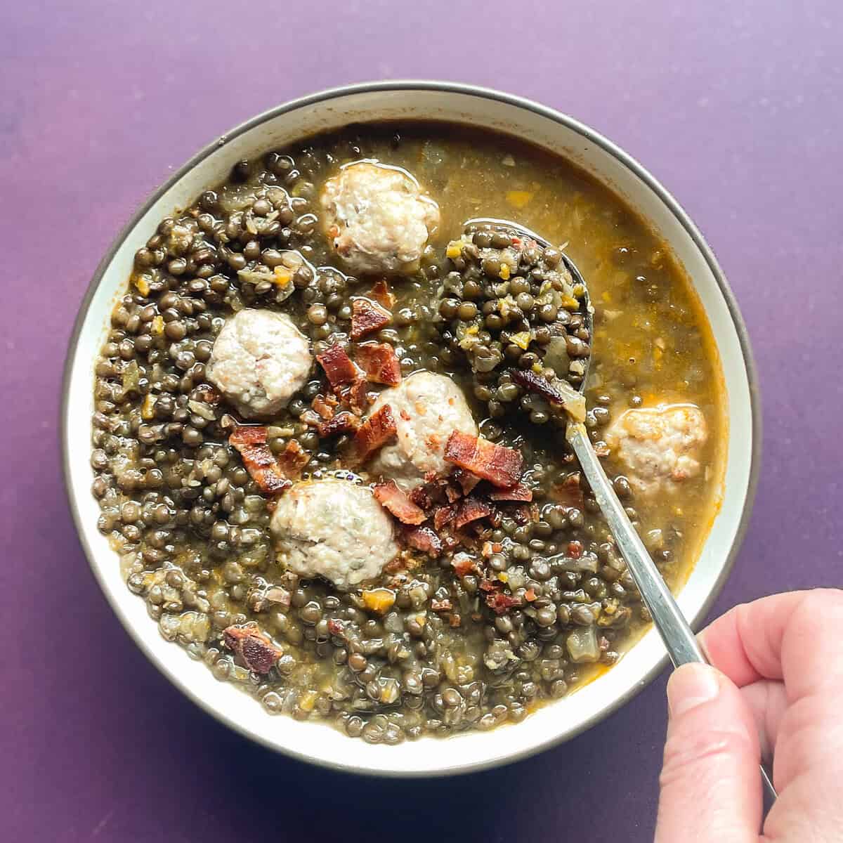 Bowl of black lentil soup topped with turkey meatballs and bacon. There is a hand holding a spoon dipping into the lentil soup.