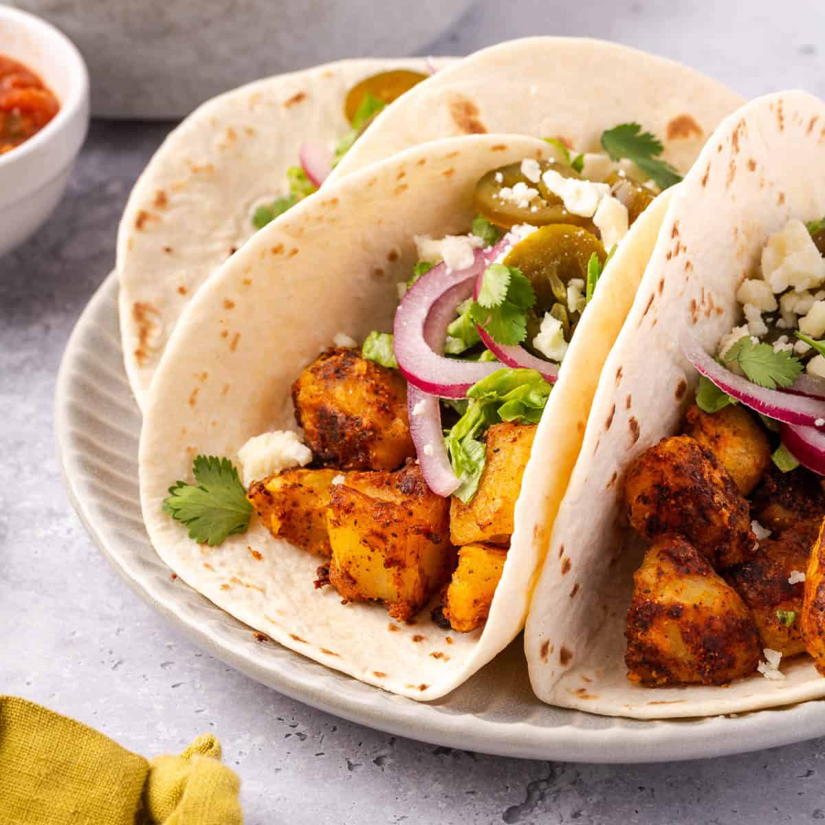 spicy potato tacos in three soft flour tortillas topped with cilantro, cheese, red onions, and other toppings.
