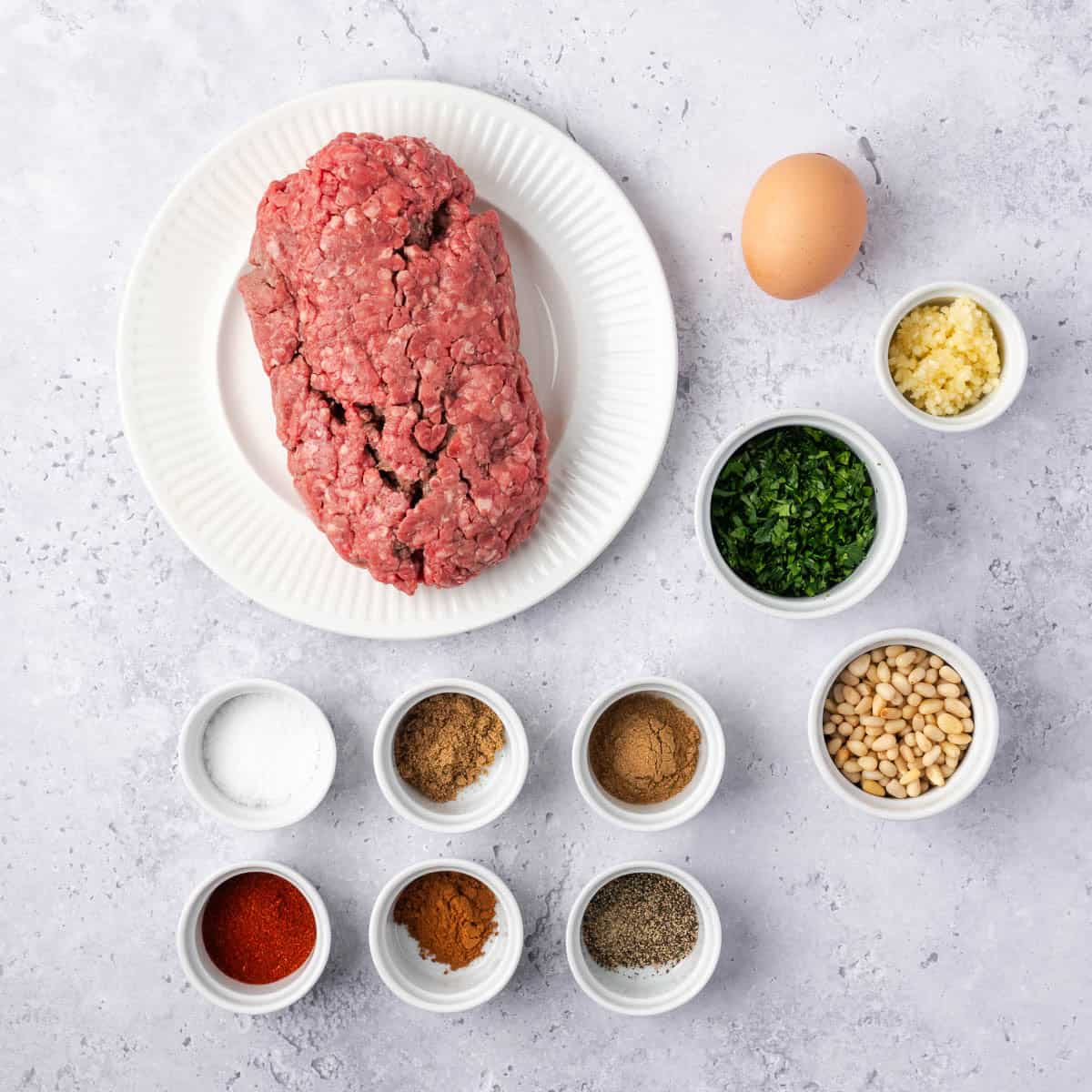 ground beef on a plate next to an egg and small white bowls filled with chopped garlic, greens, pine nuts, salt and spices.