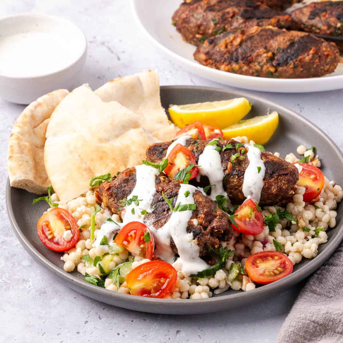 beef kofta over Israeli couscous and topped with tomatoes and cucumbers, a white sauce, chopped herbs on a plate with pita and lemon wedges.