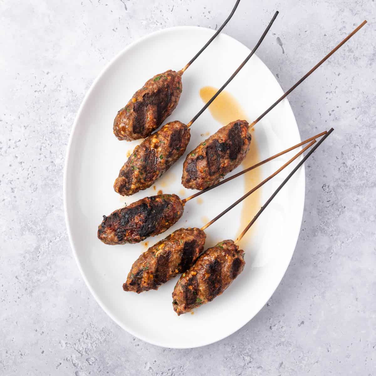 six cooked beef kofta kebabs with grill marks and still on skewers on a white platter.