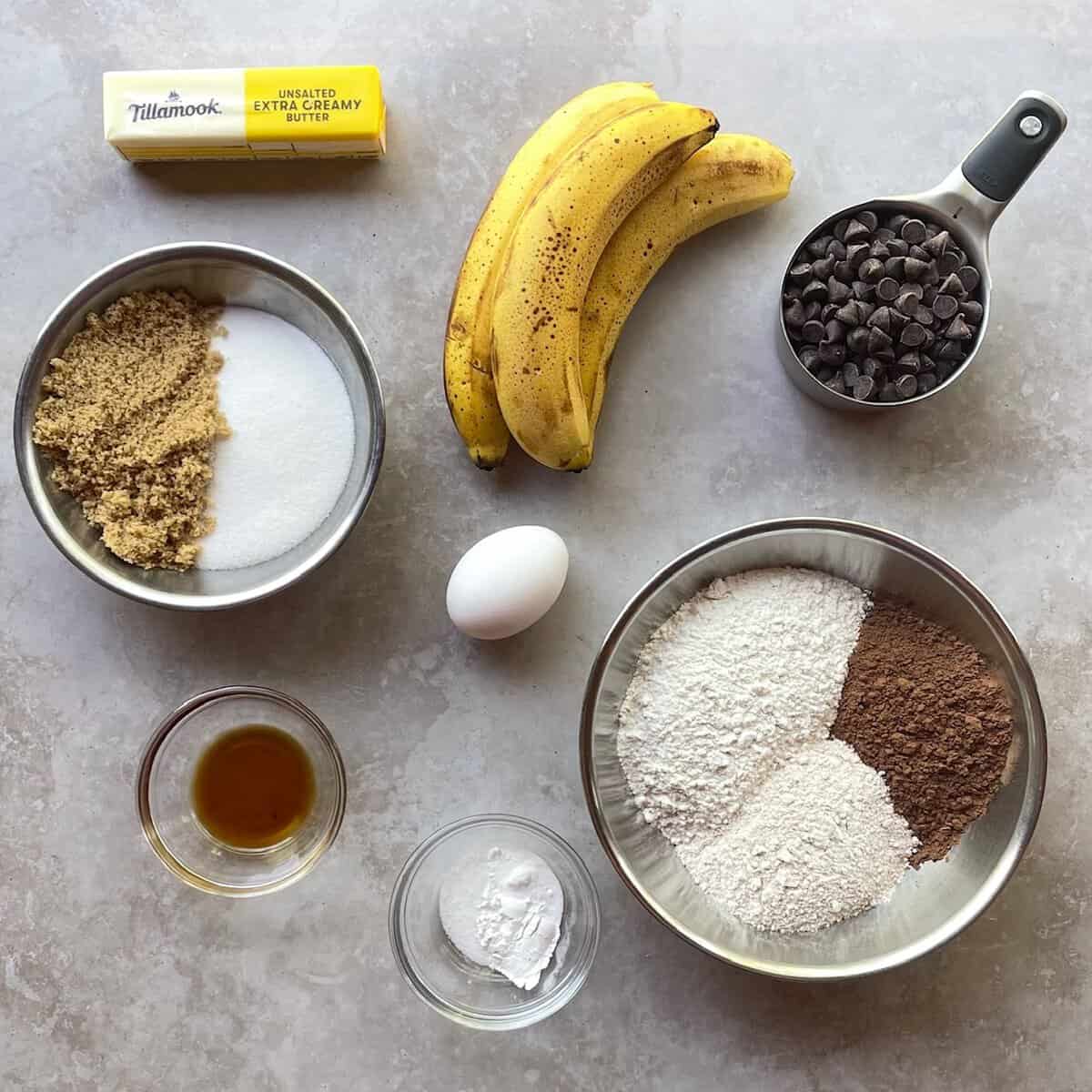 three brown speckled bananas, cup of chocolate chips, butter, brown and white sugar, cocoa and flour, egg, vanilla, and salt in small bowls.