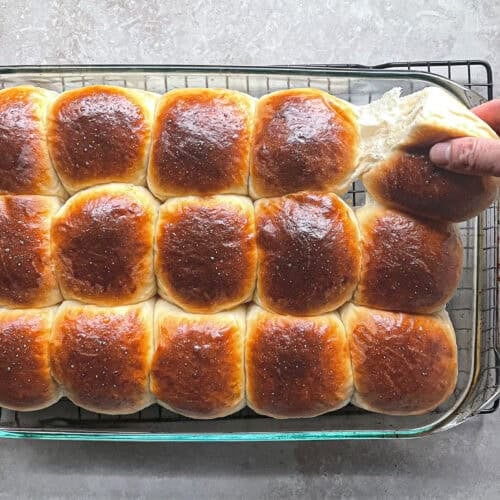 fifteen dinner rolls in a glass pan with a hand pulling away one of the corner rolls.