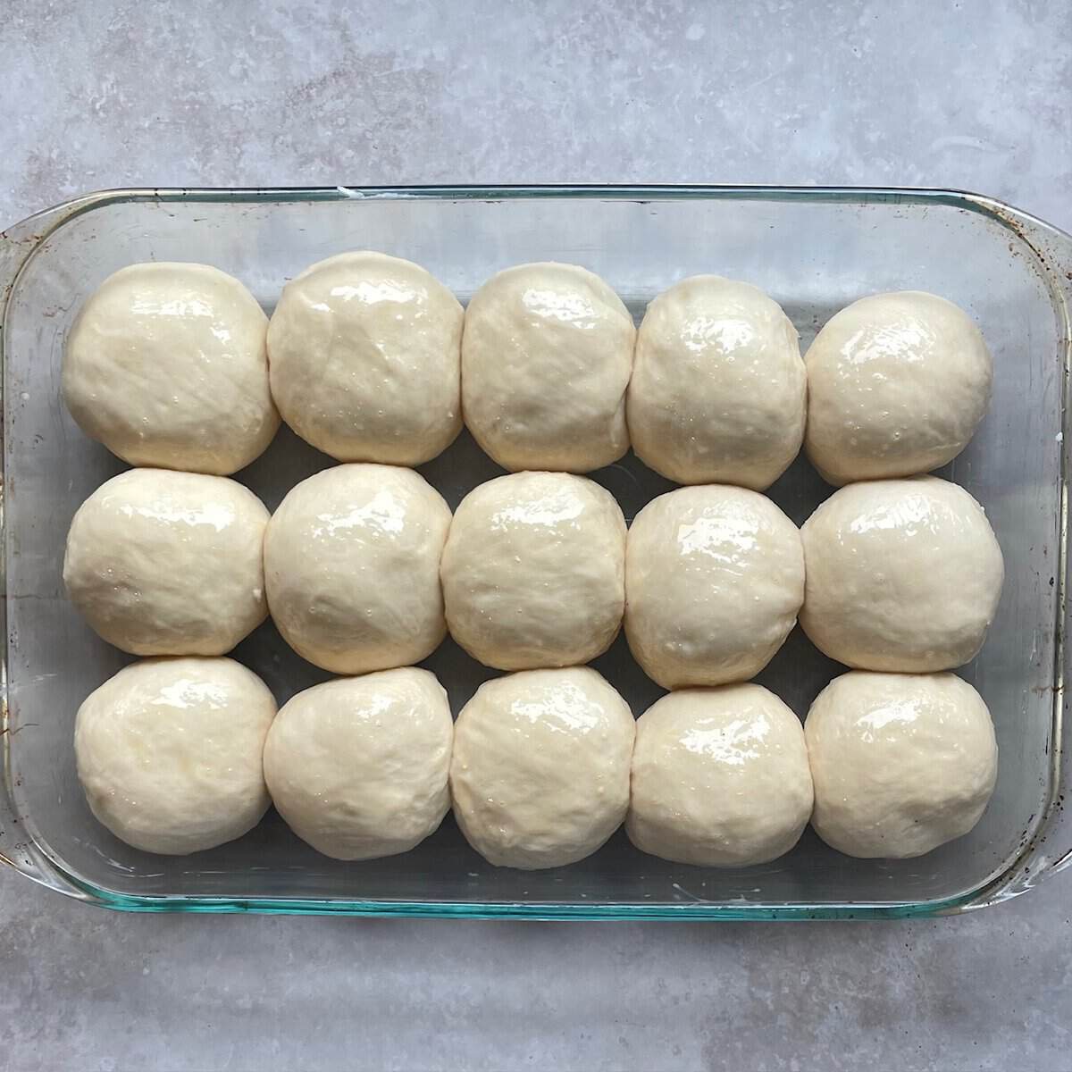 fifteen dinner rolls arranged in three rows of five rolls after rising in a glass baking dish.